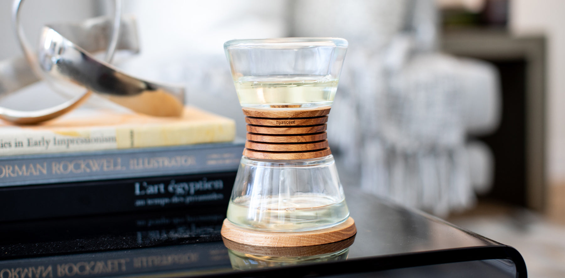 Photograph of Hyascent Fragrance Diffuser sitting on top of a black acrylic coffee table with dark decorative items in the background including books, silver sculpture, and gray sofa.